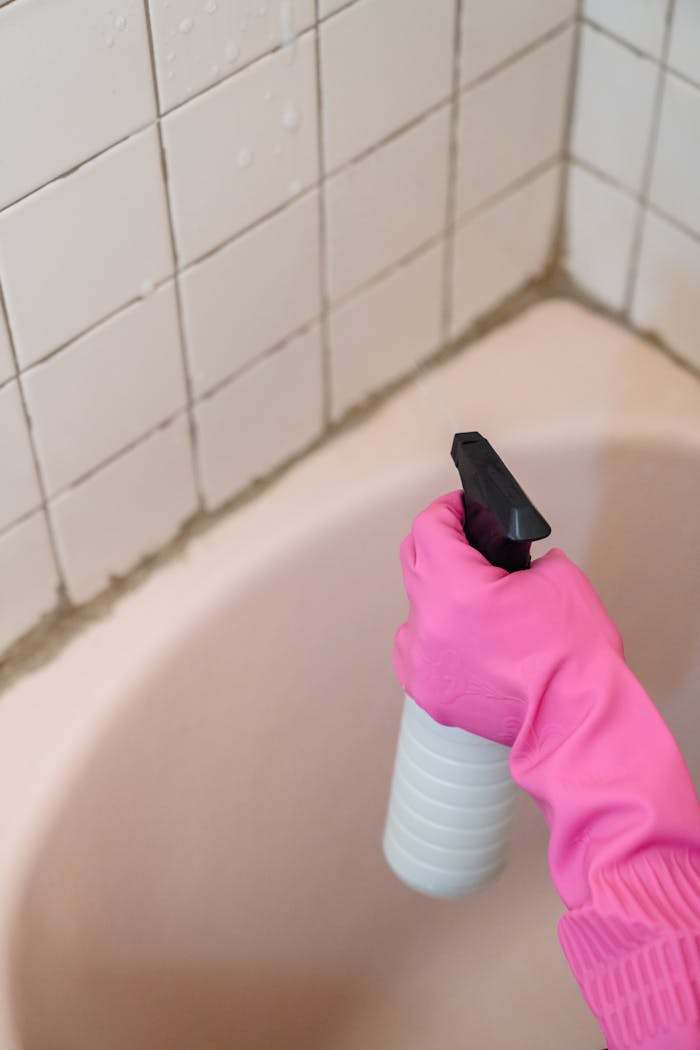 about-01 A person wearing a pink glove cleans bathroom tiles with a spray bottle.